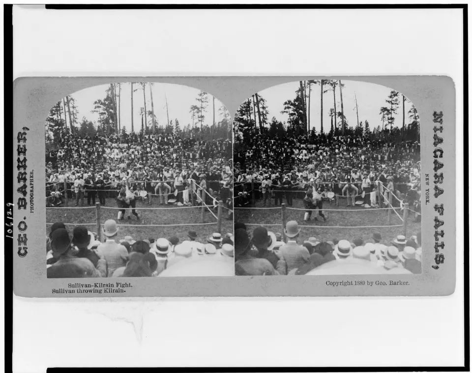 Two bare-chested boxers grappling in an outdoor ring surrounded by spectators